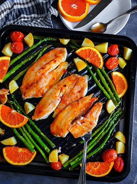 sheet pan orange ginger glazed tilapia fish with vegetables on a black oven tray