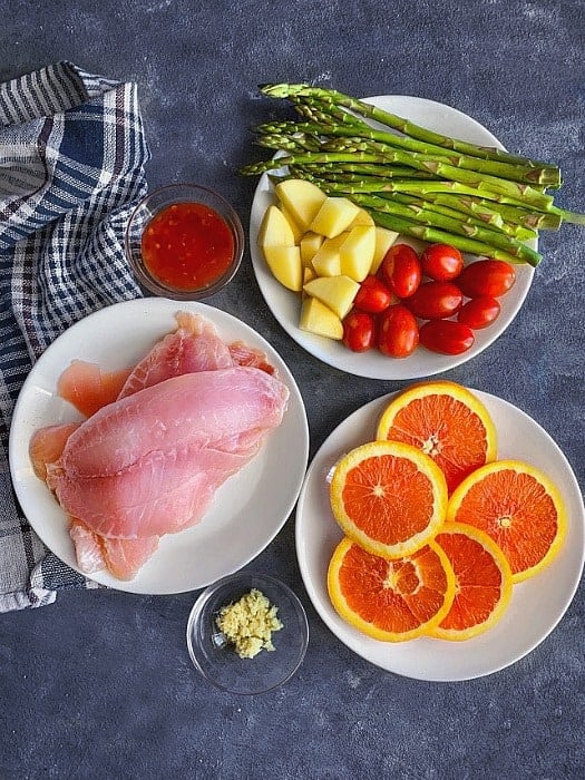 Ingredients for sheet pan orange ginger tilapia recipe. Includes cut oranges, raw tilapia fish fillet, mixed vegetables, orange and ginger marinade